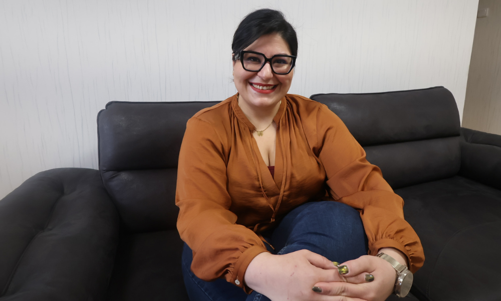 Fotoğrafta koyu renk bir koltukta oturan, gülümseyen bir kişi görülüyor. Turuncu tonlarında bir bluz, kot pantolon ve siyah çerçeveli gözlük takıyor; elleri dizlerinin üzerinde rahatça duruyor. Ortam sade, arka plan açık renkli bir duvar ve genel atmosfer sıcak ve samimi.