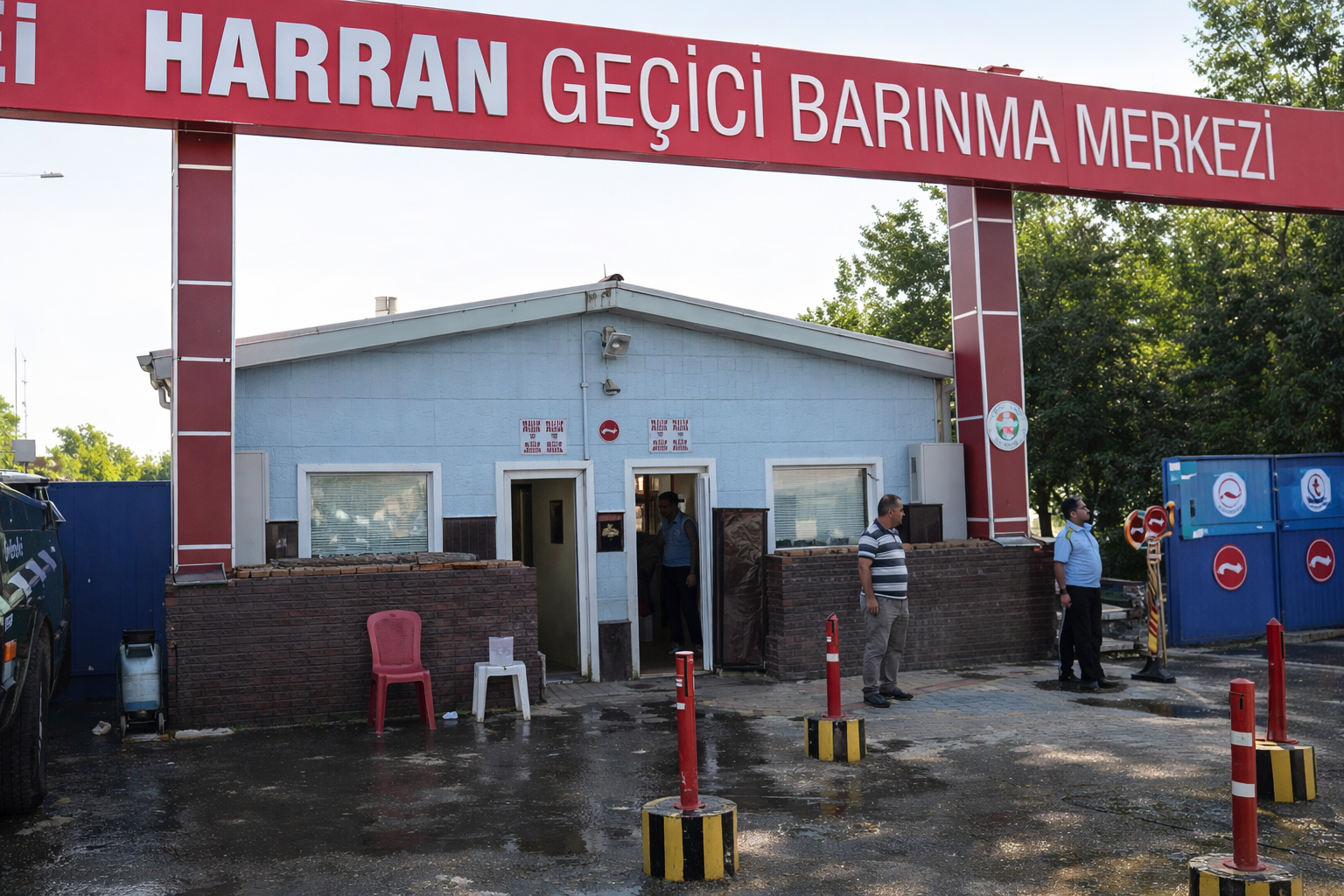 Fotoğrafta, Harran Geçici Barınma Merkezinin ana giriş kapısı görülüyor. Girişin üzerinde büyük ve dikkat çekici bir kırmızı tabela yer alıyor; üzerinde beyaz harflerle merkezin adı yazıyor. Tabelayı taşıyan geniş dikey sütunlar iki yanda yükseliyor. Alt tarafta, tek katlı, açık mavi renkte bir bina bulunuyor. Binanın ortasında ve iki yanında küçük kapılar ve pencereler var. Kapının önünde birkaç kişi ayakta duruyor; biri binanın içinden çıkar gibi, diğeri dışarıda bekler halde. Sağ tarafta iki görevli olduğu izlenimi veren kişiler göze çarpıyor. Giriş bölgesinin önünde kırmızı ve sarı çizgili güvenlik dubaları, ortalıkta ise ıslak zeminin yansıttığı ışıklar görülüyor. Sol tarafta bir araç park etmiş. Sağda ise mavi renkte, üzerinde çeşitli sembollerin bulunduğu kapılar ve bariyerler var.