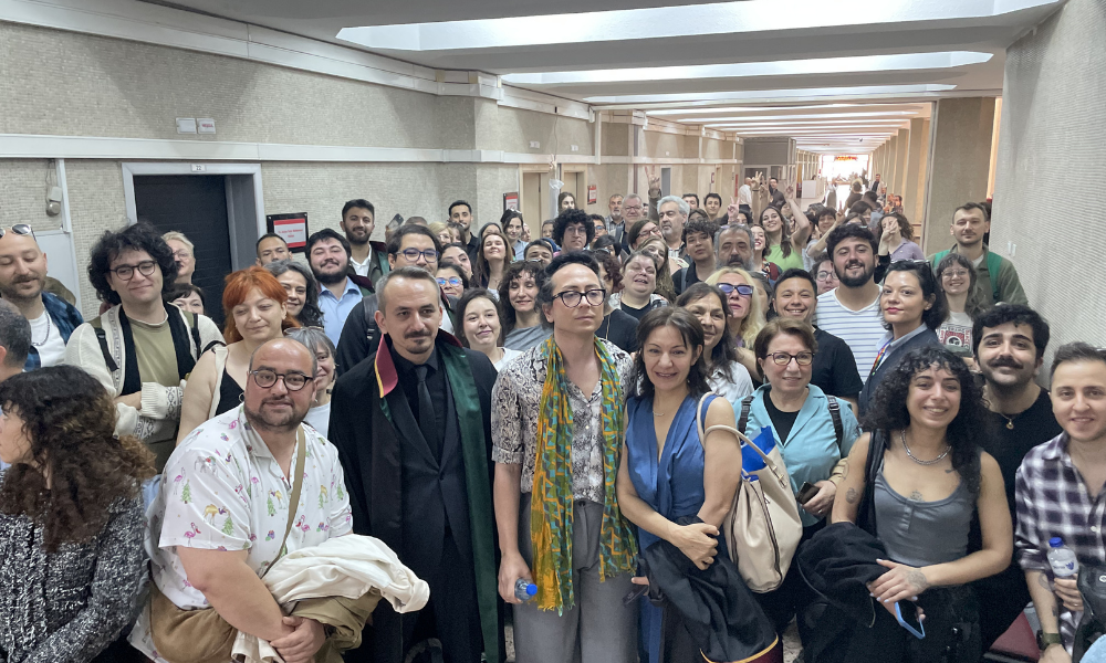 Fotoğrafta, bir adliye koridorunda bir araya gelmiş kalabalık bir grup insan görülüyor. Ön planda, üzerinde avukat cübbesi olan bir kişi ile renkli bir şal takmış olan Defne Güzel ve yanlarındaki destekçileri kameraya gülümseyerek poz veriyor. Koridorun derinliklerine doğru uzanan kalabalık, elleriyle zafer işareti yaparak beraat kararının sevincini ve dayanışmayı yansıtıyor. Mekanın yüksek tavanlı, geniş ve resmi bir devlet dairesi yapısında olması, adliye atmosferini belirgin kılıyor.