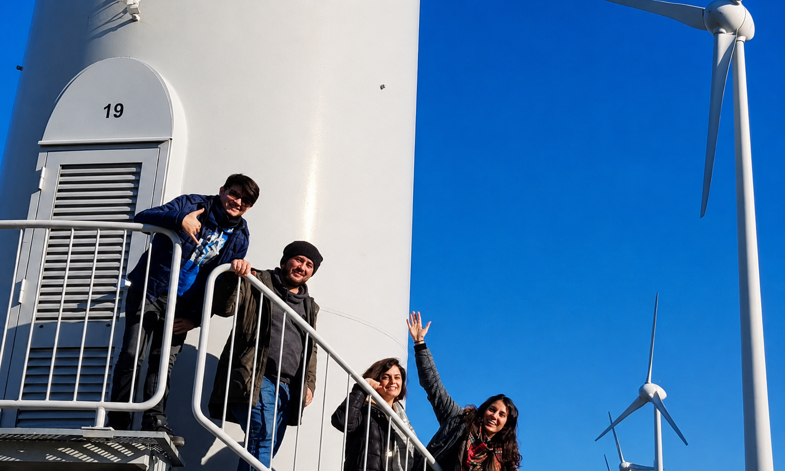 Fotoğrafta açık ve güneşli bir havada, bir rüzgâr türbininin gövdesine bağlı metal merdiven/platform üzerinde poz veren dört kişi görülüyor. Sol tarafta büyük beyaz türbin kulesi kadrajın önemli bir kısmını kaplıyor; kulenin üzerinde “19” numarası ve havalandırma panelli bir kapak bölümü var. Kişiler merdiven korkuluklarının arkasında, samimi ve neşeli bir şekilde kameraya bakıyor. Üst tarafta iki kişi, alt tarafta ise iki kişi yer alıyor; biri el sallıyor, biri başparmak işareti yapıyor. Genel duygu, bir saha ziyareti ya da yenilenebilir enerji tesisinde çekilmiş anı fotoğrafı izlenimi veriyor. Sağ tarafta berrak mavi gökyüzü geniş bir boşluk oluşturuyor ve arka planda iki rüzgâr türbini daha görünüyor. Fotoğrafın kompozisyonunda beyaz türbin gövdesi, gri metal korkuluklar ve yoğun mavi gökyüzü arasında güçlü bir kontrast var. Işık sert ve doğrudan geldiği için gölgeler belirgin; bu da görüntüye net, canlı ve açık hava belgeseli hissi katıyor.