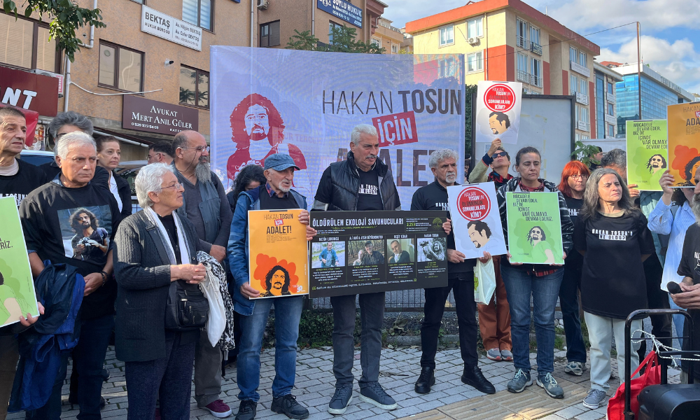 Gündüz saatlerinde şehir merkezindeki bir cadde kenarında toplanan kalabalık bir grup, üzerinde Hakan Tosun’un çizimi ile büyük harflerle "HAKAN TOSUN İÇİN ADALET" yazılı beyaz pankartın arkasında yan yana duruyor. Pankartın önünde ve çevresinde yer alan kadın ve erkekler, ellerinde Tosun’un fotoğraflarının, "Öldürülen Ekoloji Savunucuları" listesinin ve adalet talebini belirten çeşitli dövizlerin yer aldığı sarı, yeşil ve beyaz renkli afişler taşıyor. Katılımcıların çoğu siyah ve koyu renkli kıyafetler giymiş olup bazılarının tişörtlerinde Tosun’un fotoğrafı veya ismi yer alıyor. Grubun arkasında avukatlık ve sigorta ofislerinin tabelalarının bulunduğu çok katlı şehir binaları ile açık, mavi bir gökyüzü görülüyor.